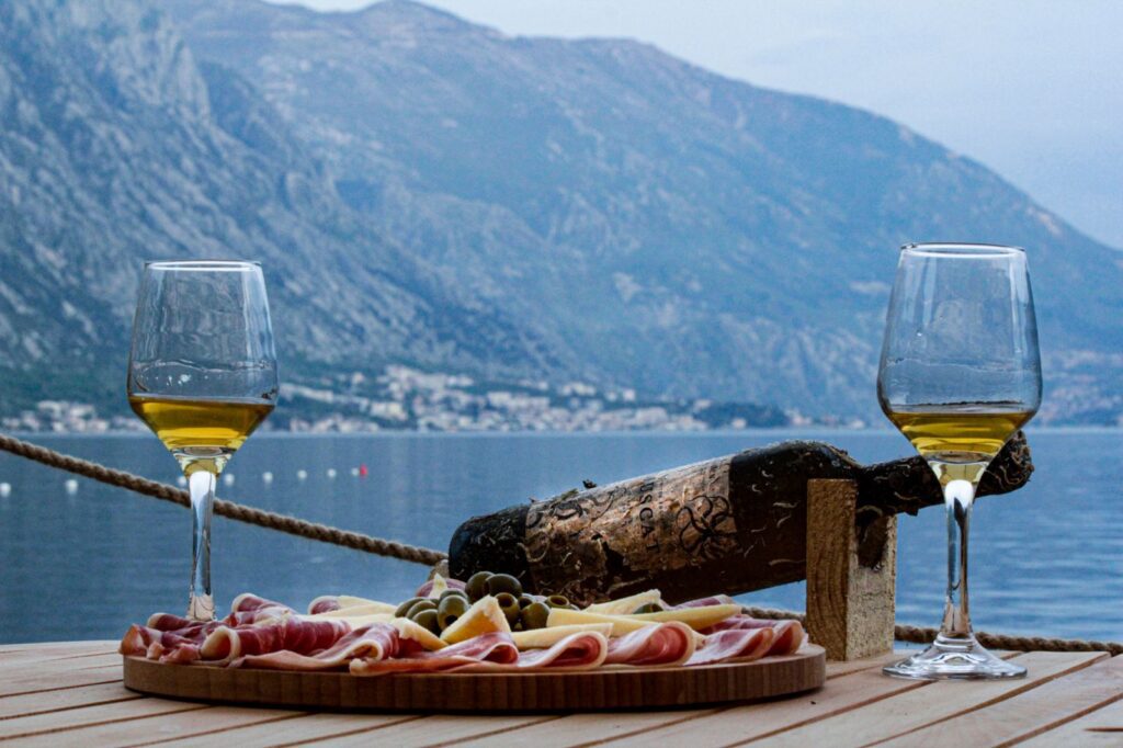 kotor boat tours: wine tasting tour kotor: image of a plate with prosciutto and two glasses of wine and a wine bottle, hills and sea in the background, noon